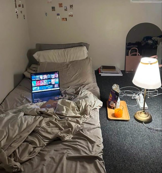 A cozy bedroom scene with an unmade bed featuring gray bedding, a laptop open to a streaming service, a small lamp, snacks on a tray, and polaroid photos on the wall.