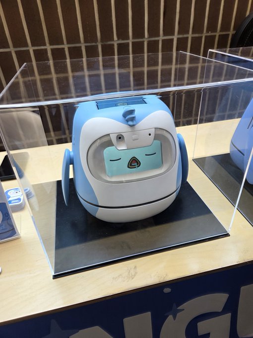 A blue and white robotic toy with a light blue face featuring closed eyes is displayed inside a clear acrylic case on a counter.