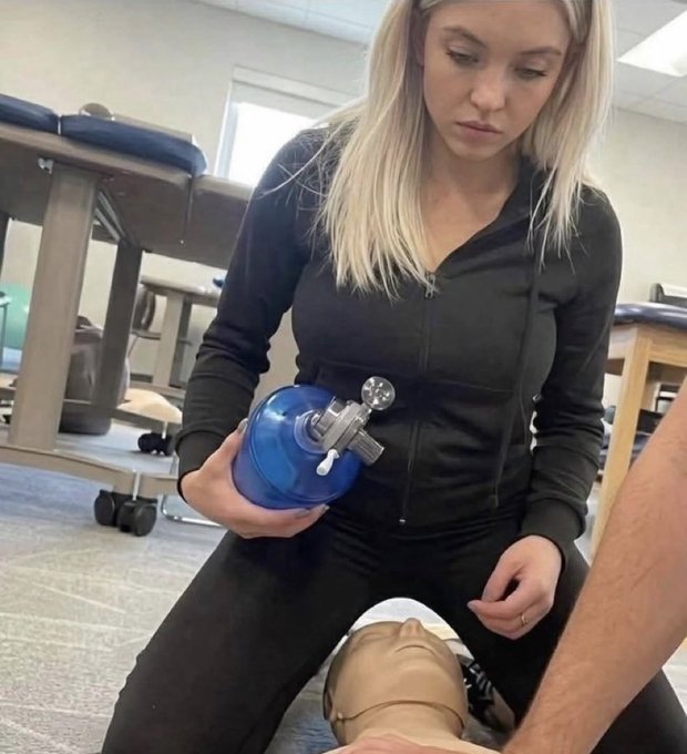 A blonde woman in a black tracksuit holds a blue resuscitation bag (Ambu bag) while practicing on a medical training mannequin, illustrating the "It is actually fine." caption by showing her calmly managing the equipment during a CPR or airway training scenario.