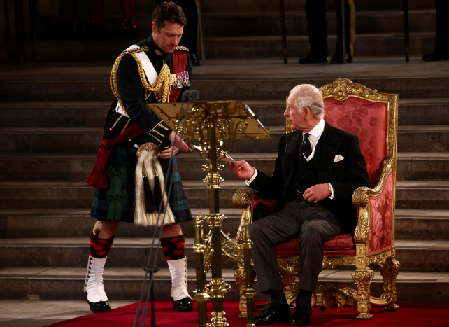 A man in formal military dress, including a kilt and medals, stands beside an ornately carved red and gold throne where King Charles III sits holding a pen, likely during the State Opening of Parliament. The post reflects concern over the departure of trusted courtiers, expressing hope that the King remains well-supported amid rumors of political turbulence.