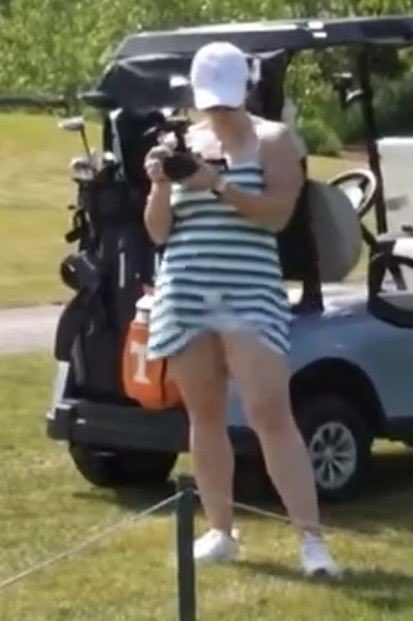A woman wearing a white cap and a black and white striped dress stands on a golf course, looking down at her phone, with a golf cart filled with clubs parked behind her.