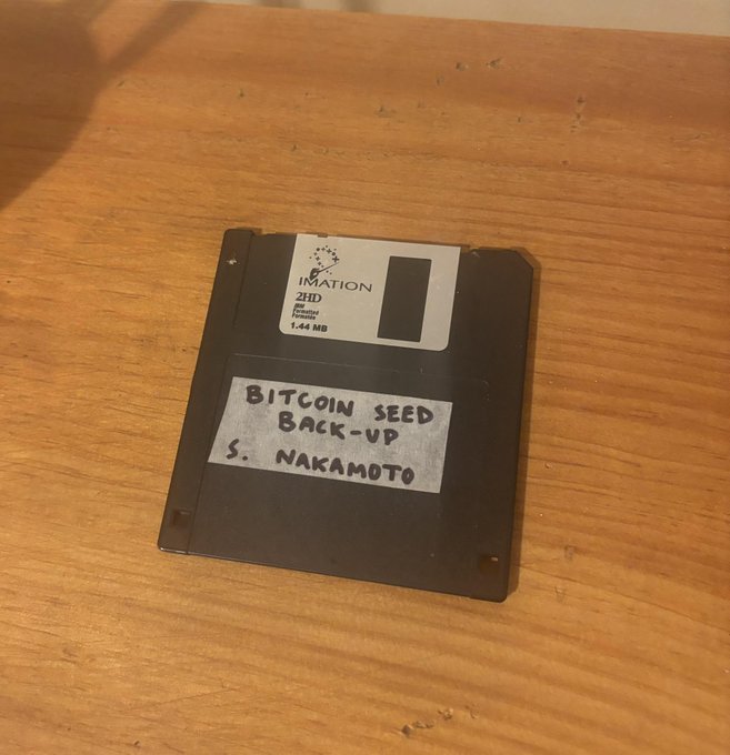 The image shows an old floppy disk with the words "BITCOIN SEED BACK-UP S. Nakamoto" written on it, indicating that someone has repurposed this computer junk for a new use. The floppy disk appears to be in good condition and is placed on top of a wooden table or desk.