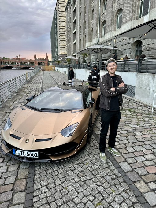 The image features a man standing next to a gold sports car, posing for a photo. The car has a license plate that reads "B6T 661". In addition to the man and the car, there are two umbrellas visible in the scene - one near the center of the image and another towards the right side. There is also an umbrella located on the left side of the image.