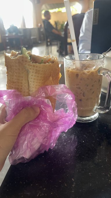 A first-person view of a hand holding a crepe wrapped in a purple plastic bag next to a glass mug of iced coffee, taken in what appears to be a cafe setting.