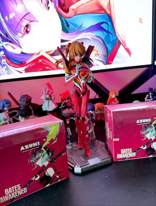 An Asuka Langley figure from 'Gates Awakening' stands between two 'AZUKI' branded boxes, with anime artwork displayed on a screen behind the display.
