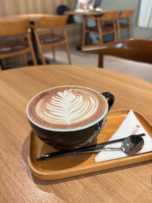The image features a cup of coffee sitting on top of a wooden tray, accompanied by silverware including a spoon and fork. The background shows several chairs and tables arranged around dining tables, suggesting that this scene takes place in a restaurant or cafe setting. The caption reads "Gm CT What's the move today legends? Jump into @KGeN_IO Campaign (ends 13/6) #clear daily quests on @mamebnb #finish #HerondBrowser Cup quests #share some alpha & collab with new projects".