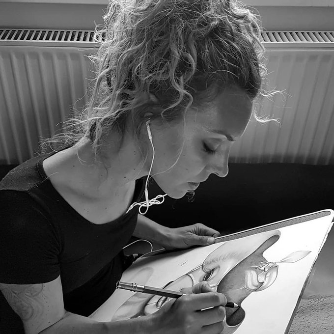 A black and white photograph of a curly-haired woman wearing earbuds, intently sketching a detailed figure in a large notebook with a mechanical pencil.