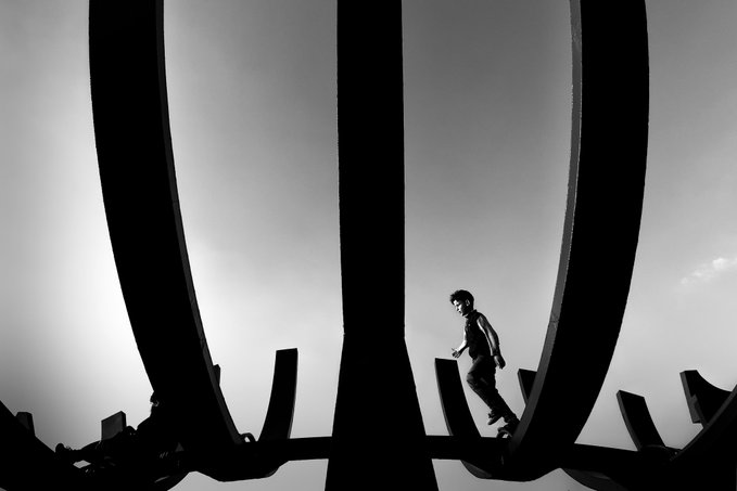 A dramatic black and white silhouette photograph showing a person walking across the curved metal ribs of a large architectural structure against a bright sky.