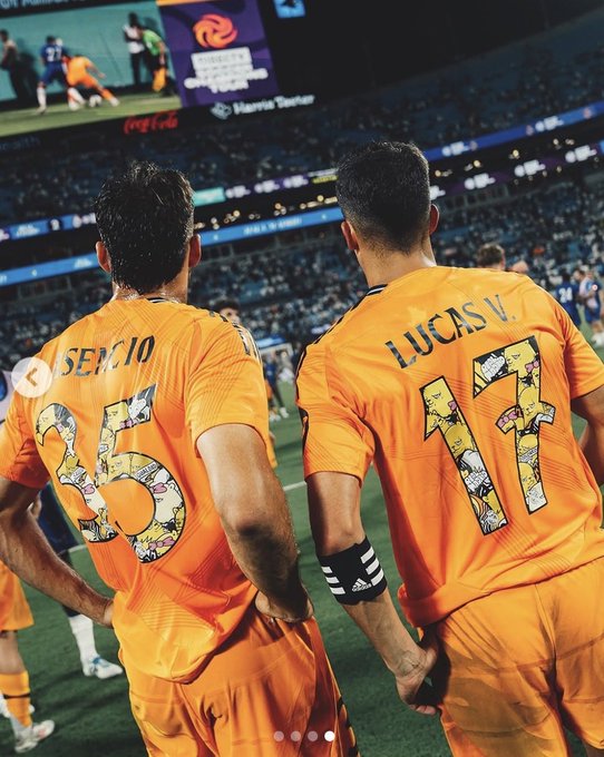 A rear view of two soccer players in orange Los Angeles Galaxy jerseys with the names 'SERCIO 5' and 'LUCAS V. 17', standing on the field with a stadium crowd in the background.