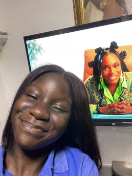 A selfie of a smiling young woman in a blue shirt, with a television in the background displaying an image of a young girl with green hair ties.