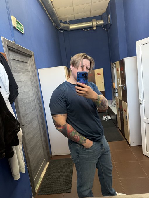 A man with blonde hair takes a mirror selfie with a blue smartphone in a hallway with blue walls, wearing a gray t-shirt and blue jeans that reveal tattooed arms.
