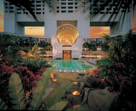An ornate indoor atrium features a swimming pool surrounded by lush tropical plants, lounge chairs, and a grand domed ceiling illuminated by warm sunset light.