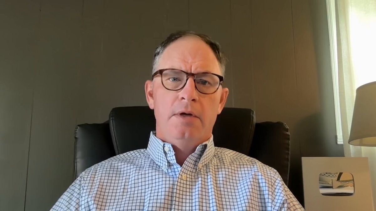 The image shows a man wearing glasses sitting in an office chair, with his face turned towards the camera. He appears to be giving a presentation or participating in a video call. The background features a wall that has a window, suggesting that he is likely indoors. There are also two lamps visible in the room, one on each side of the man's chair.