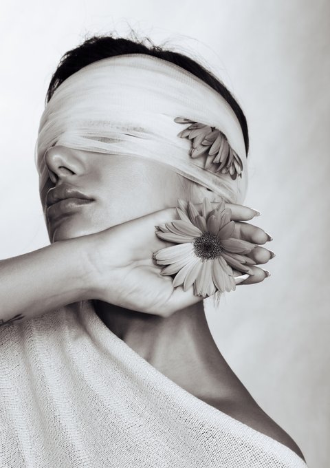 This black and white photo features a woman with two flowers placed on either side of her face, which appears to be tied to an unseen object or person's head using a scarf. She is wearing a white shirt that contrasts with the overall monochrome atmosphere of the image. The image has a post-processing effect applied to it, giving it a vintage appearance and adding a layer of artistic depth to what could otherwise have been just another ordinary photo capturing a moment in someone's life.