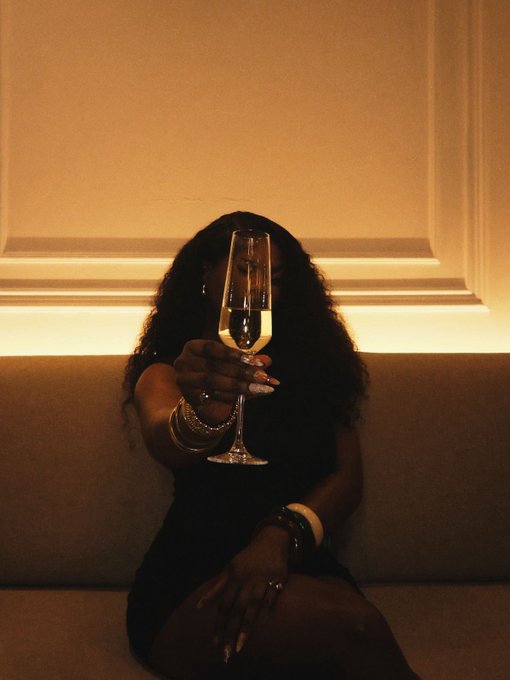 A woman seated on a beige couch holds up a champagne glass, obscuring her face in the warm-lit setting, evoking a celebratory toast to new beginnings and the hopeful sentiment, "May the odds be in our favour."