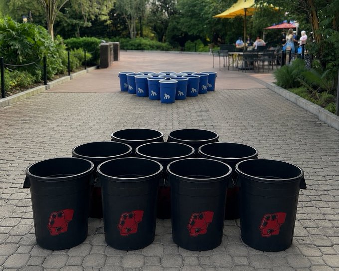Black buckets with a red logo and blue cups with a white logo are arranged on a brick patio in an outdoor park setting.