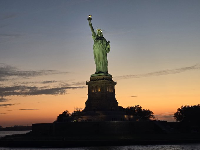 The Statue of Liberty illuminated against a sunset sky with orange and blue gradients, viewed from the water.