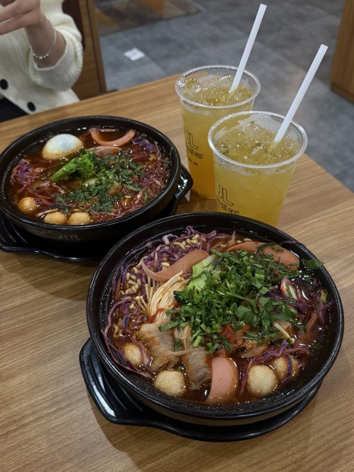 Two bowls of spicy noodle soup topped with herbs, vegetables, and meat sit on a wooden table alongside two plastic cups of iced orange drinks.