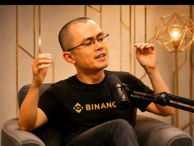 The image features a man sitting in a chair, wearing glasses and a black t-shirt with the word "BINANC" printed on it. He appears to be engaged in conversation or giving a speech, as indicated by his hand gestures. The background of the image is white, which contrasts nicely with the gold accents around him. There are two additional objects in the scene: a lamp and a geometric sculpture that adds an artistic touch to the setting.