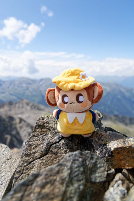 A small plush monkey toy wearing a yellow hat and outfit sits on a rocky mountaintop against a blue sky.
