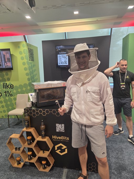 A smiling man wearing a beekeeper's hat and white protective suit poses at a trade show booth for 'HiveBits' with honeycomb-shaped product displays.