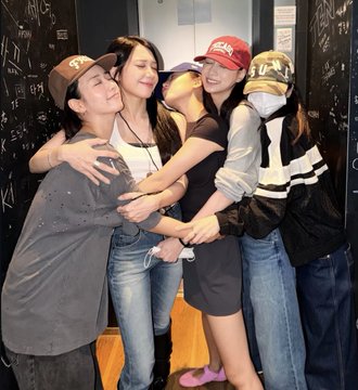 A group of five friends share a warm group hug in front of a blackboard covered in chalk writing, with one person wearing a red "Yankees" cap and another wearing a face mask. The "Apingoo family" caption reflects the close-knit, familial bond visible in their joyful embrace and playful attire.