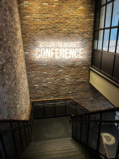 An interior stairwell with brick walls and a neon sign that reads 'BLEEDIN' TREASURES CONFERENCE'.