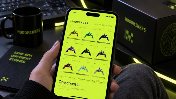 A hand holds a smartphone displaying a bright yellow "HOODMINERS" NFT mint page with text "One chassis" and "WORLDHOOD CHAIN," surrounded by branded merchandise including a mug and box.