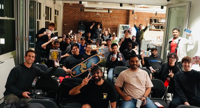A group photo of around twenty people gathered in a loft-style room with brick walls, holding up cards and a skateboard while celebrating enthusiastically.