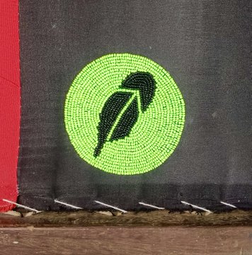 A close-up of a circular beaded patch with a black feather design on a bright green background, stitched onto dark fabric next to a red section.