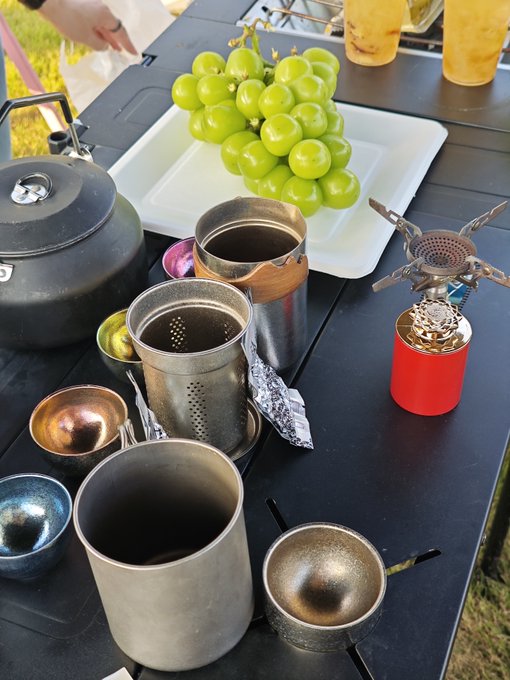 A kettle, camping stove, cups, and a tray of fresh green grapes are set up on an outdoor table, suggesting someone is enjoying a casual outdoor brewing or tea session. The post's playful tone about reporting your activities with a photo fits as the user shares this snapshot of their relaxed outdoor setup. #报备