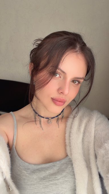 A selfie of a young woman with brown hair, a nose piercing, wearing a gray tank top and an embellished choker necklace, posing in front of a textured wall.