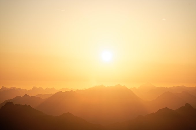 A serene landscape photograph captures a bright golden sunrise or sunset over layers of silhouetted mountain ridges fading into a warm, hazy orange atmosphere.