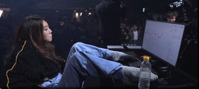 A woman with long dark hair relaxes with her feet up, looking at a computer monitor displaying stock charts in what appears to be a trading event setting.