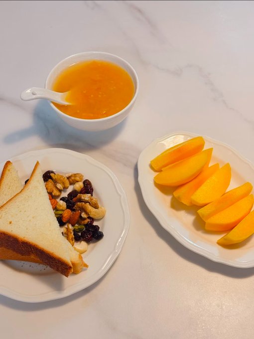 A meal laid out on a white marble surface, featuring a bowl of orange soup, a plate with a sandwich and mixed nuts/dried fruit, and a plate of sliced mango.