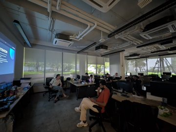 People working at computers in a modern open-plan office with exposed ceilings and large windows.