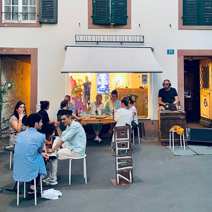 A group of people gather around a table outside a wine bar at street number 21, with a DJ wearing headphones performing on a wooden crate to the right.