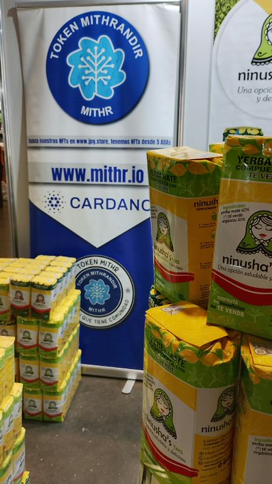 A trade show booth displays banners for 'TOKEN MITHRANDIR (MITHR)' and 'CARDANO' alongside stacked packages of 'ninusha's' yerba mate and tea products, with text indicating the token is tradable on www.jpgs.store since August 5.
