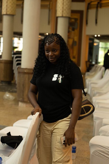 A young woman smiles warmly at the camera while holding a draped chair and wearing a black t-shirt with the "SX" Solana logo at the Solana Summit Nigeria Day 2 event.

The image relates to the post as a candid photograph captured by @lifeofedi_ during the Solana Summit Nigeria, showcasing an attendee wearing official Solana-branded merchandise at the conference venue.
