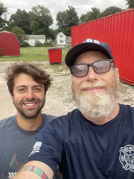 A selfie of two men smiling outdoors in front of red metal buildings, with one wearing a gray shirt and the other wearing a navy 'IRONDALE FIRE' shirt and a 'FD' cap.