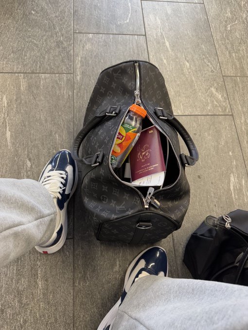 An open Louis Vuitton monogram duffle bag sits on a tiled floor, revealing various items inside including what appears to be a beverage bottle and a burgundy container, with the photographer's white and navy sneakers visible.