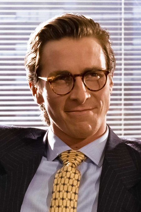 A man with slicked-back brown hair, round tortoiseshell glasses, and a pinstripe suit with a patterned gold tie smiles slightly in an office setting with window blinds behind him. The image appears to illustrate the Ava Labs leadership announcement, though it does not depict the named executives Charley Cooper, John Wu, or Lydia Chiu.