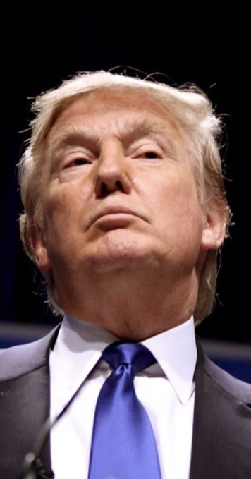 Close-up portrait of Donald Trump wearing a dark suit, white shirt, and blue tie, photographed from a low angle against a dark background.