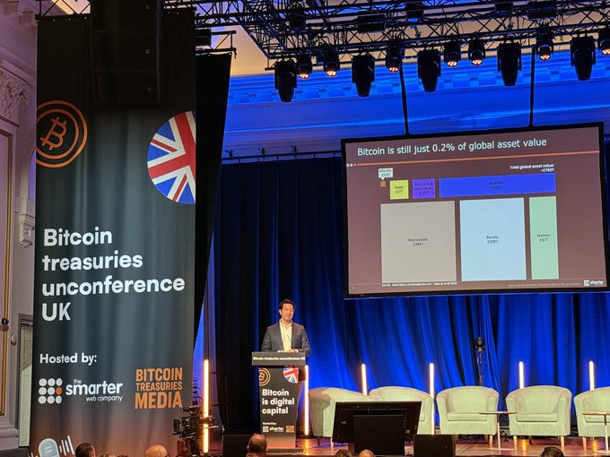 In the image, a man is standing at a podium with a large screen behind him displaying a map of global wealth. The man appears to be giving a presentation or speech related to Bitcoin and its impact on the economy. The audience consists of several people seated in chairs around the stage, attentively listening to the speaker's words.