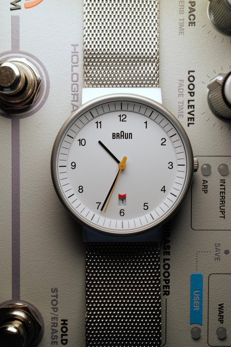 A Braun wristwatch with a mesh band rests on a Hologram Effects pedal labeled 'Stop/Erase Hold' and 'Loop Level,' showing orange hands pointing to about 7:32 with a date window at 6.