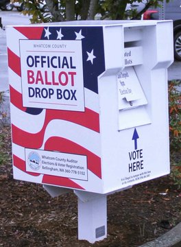 An official "Whatcom County" ballot drop box decorated in red, white, and blue American flag colors stands ready for voters to cast their ballots, with a "VOTE HERE" label and an arrow indicating where to insert ballots. The image of secure ballot drop boxes relates to the post by symbolizing the voting process that crypto lobbying group Fairshake aims to influence with its $30 million campaign targeting former Senator Sherrod Brown in Ohio.