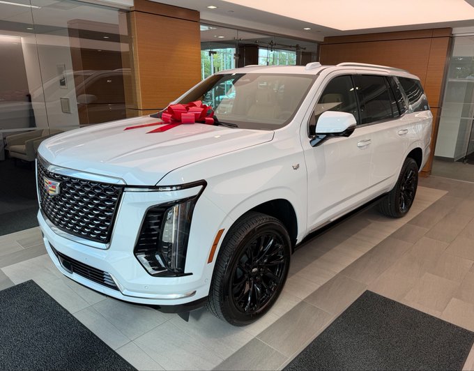 A pristine white Cadillac Escalade, adorned with a festive red bow on its hood, sits prominently in what appears to be a luxury dealership showroom, likely recently delivered. The image accompanies a post celebrating a 9th addition to a personal collection, humorously dubbed "the money glitch."