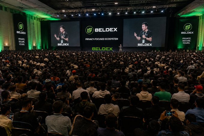 A large conference event for BELDEX, with a speaker presenting on stage in front of massive screens displaying 'BELDEX - PRIVACY-FOCUSED ECOSYSTEM' to an audience of thousands.