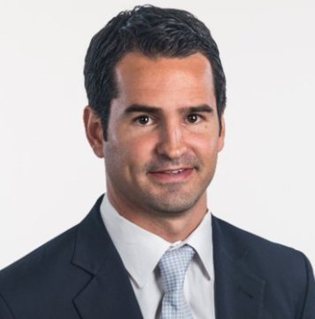 A professional headshot of a smiling middle-aged man with short dark hair wearing a dark suit, white shirt, and light blue patterned tie against a white background.