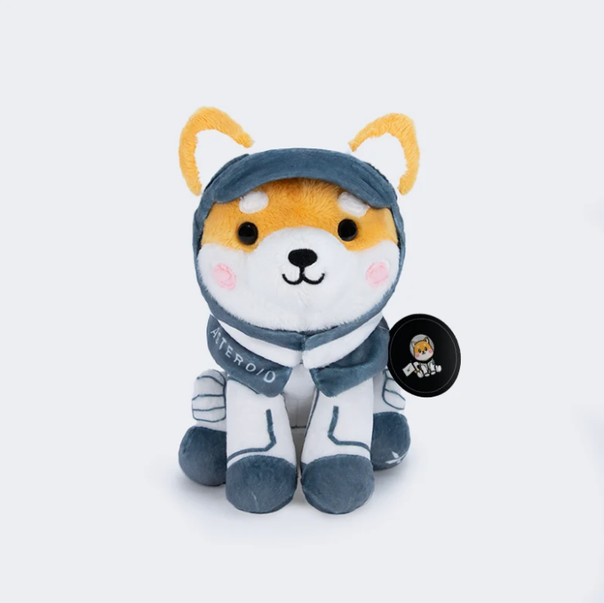 A plush toy of the Android mascot dressed in an astronaut suit, featuring a dog-like face with orange ears, accompanied by a small Android pin.